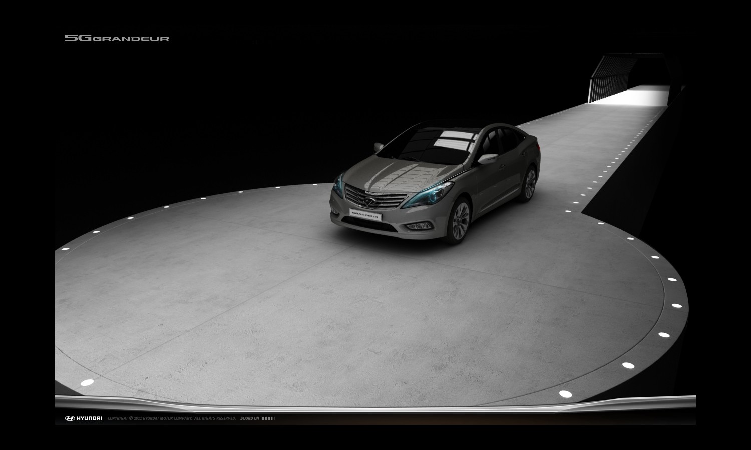 어두운 배경의 실내 공간에 회색 그랜저 HG 차량이 비스듬히 세워져 있다. 전면 그릴과 헤드램프가 강조되어 있으며, 바닥과 차량 표면에 조명이 반사되어 고급스러운 분위기를 연출한다.