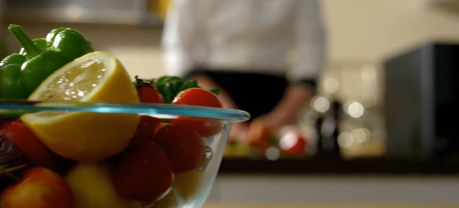 유리 볼에 담긴 방울토마토와 채소가 놓여 있고 뒤쪽에서 요리하는 모습